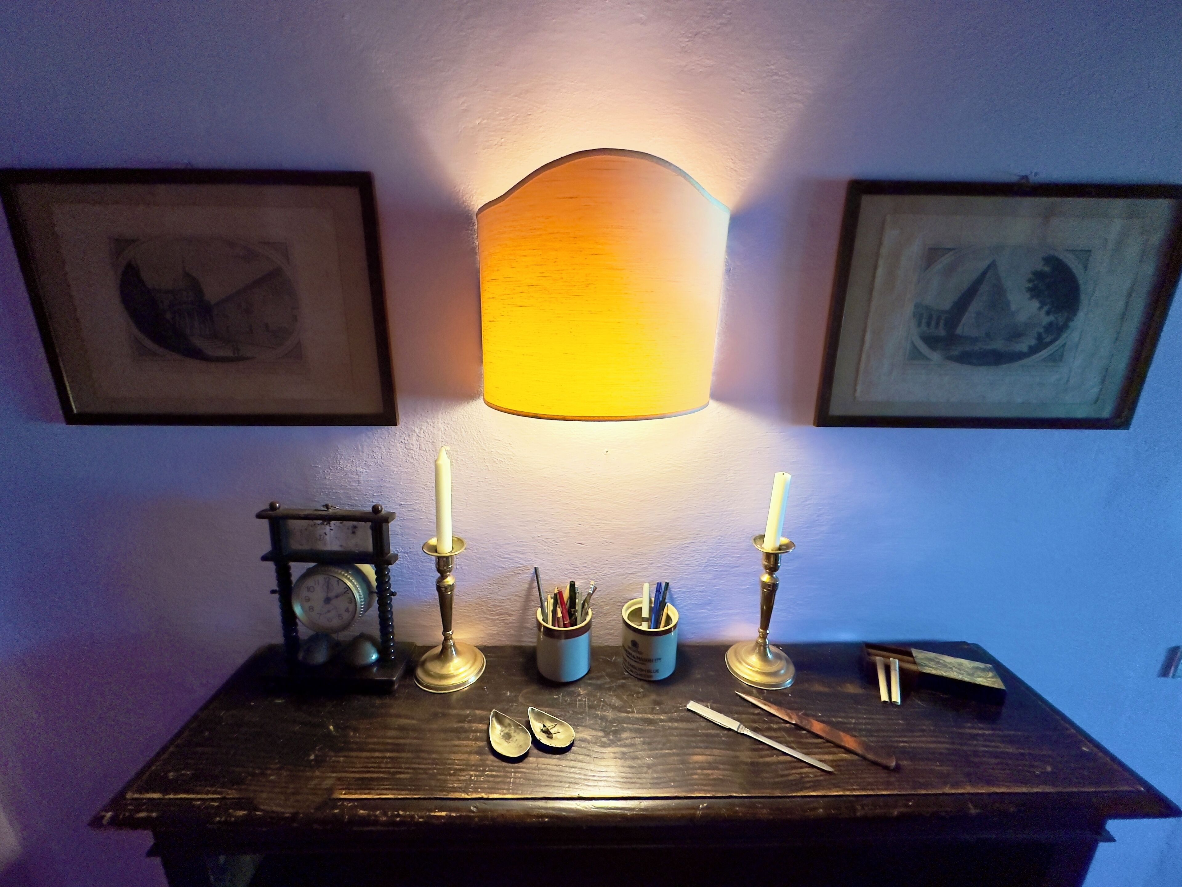 A wooden table with a lamp, two candles, a clock, and a framed picture.