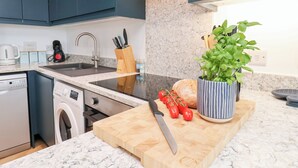 A modern kitchen with dark blue cabinets, a stainless steel sink, a Bosch dishwasher, a washing machine, a wooden cutting board, a knife, cherry tomatoes, a basil plant in a striped pot, and a coffee maker. - Belle's View (Scarborough)