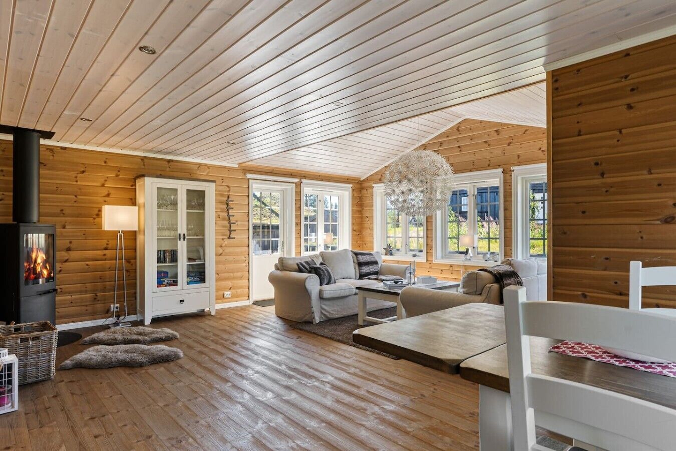A cozy cabin interior with a wood-paneled ceiling, a fireplace, a sofa, and a dining table.