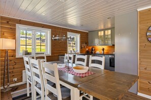 A dining area with a wooden table and chairs, a kitchen with grey cabinets, and a window with a view of greenery.