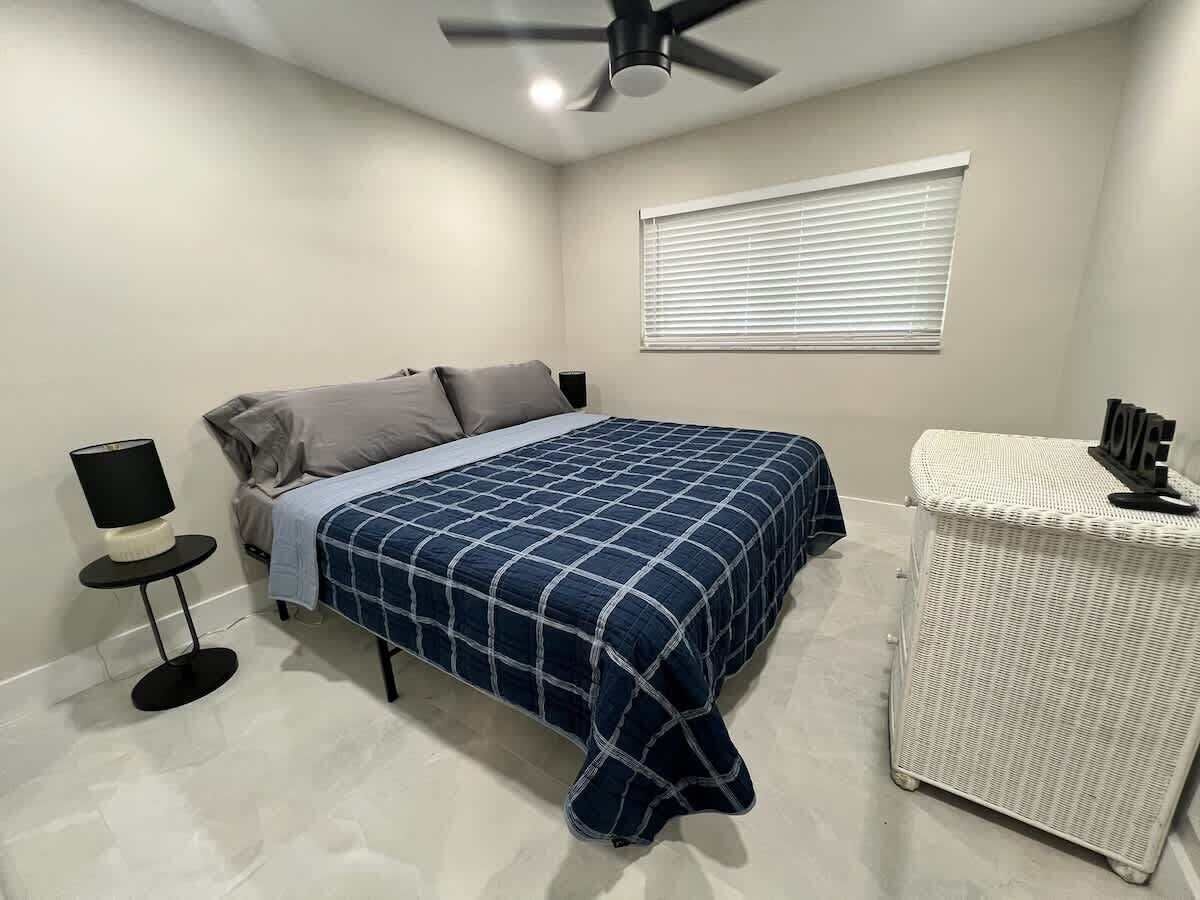 Un dormitorio con cama, una mesita de noche con lámpara, ventilador de techo y ventana con persianas.