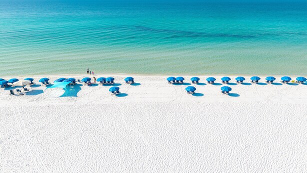 Een strand met wit zand en helderblauw water. Er staan verschillende blauwe parasols en ligstoelen langs de kust.