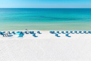 Une plage de sable blanc bordée d’une eau d’un bleu cristallin. Plusieurs parasols bleus et des chaises longues sont disposés le long du rivage.
