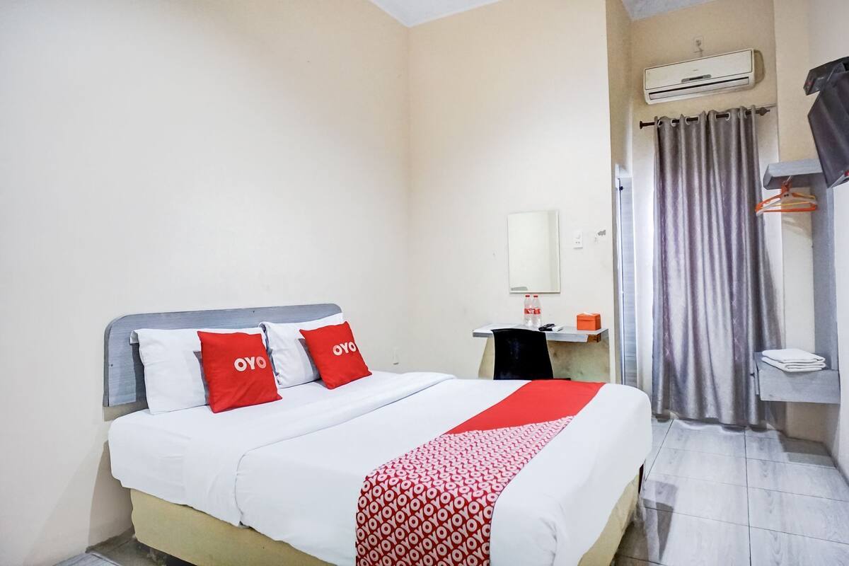 A hotel room with a bed, a bedside table, an air conditioning unit, a mirror, and a curtain.
