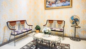 Een kamer met twee rieten stoelen, een bijzettafeltje met een bloemenvaas, een ventilator en een schilderij van paarden aan de muur.