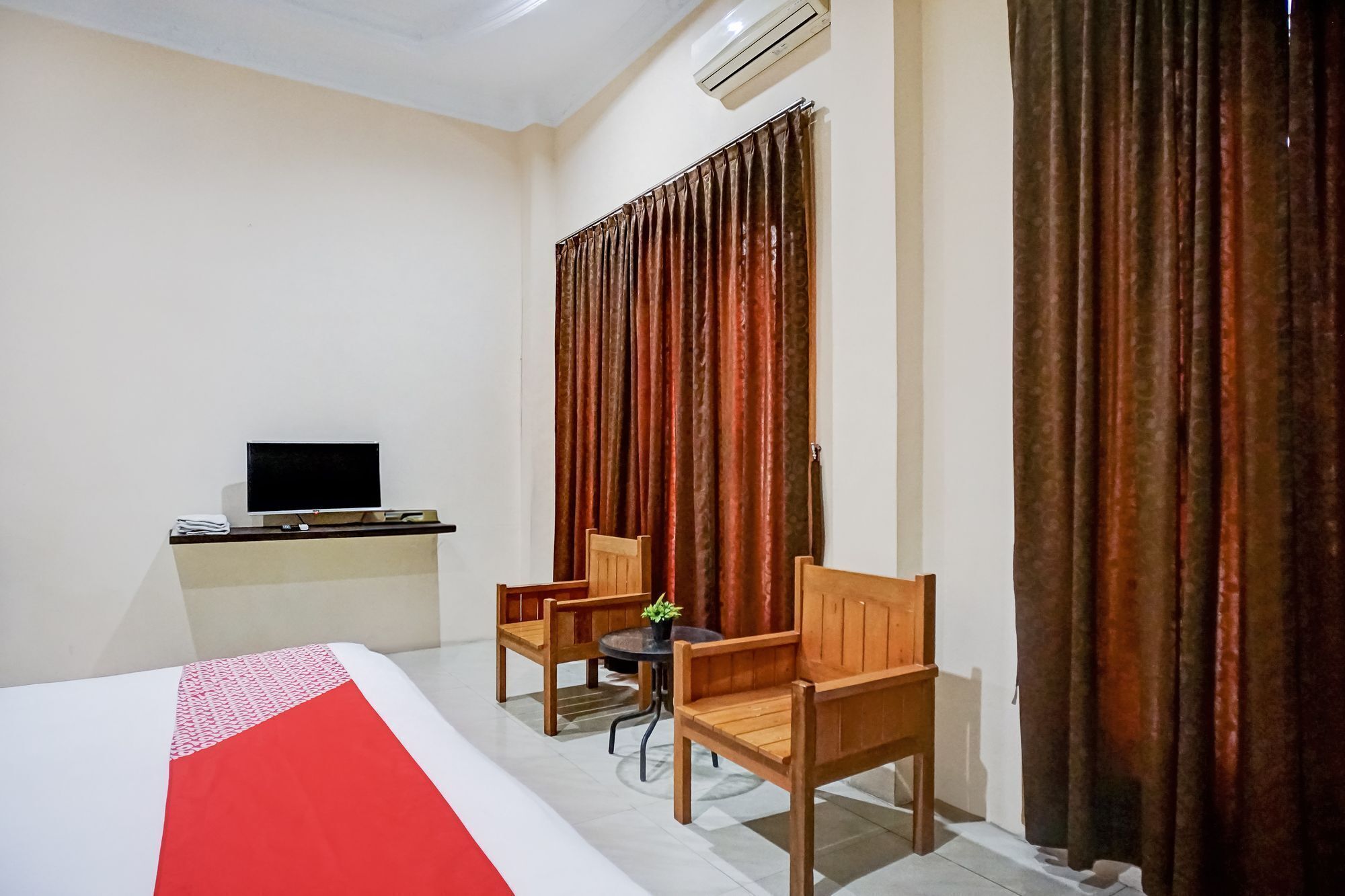 A hotel room with a bed, a desk with a computer, two wooden chairs, a small table with a plant, and a window with curtains.