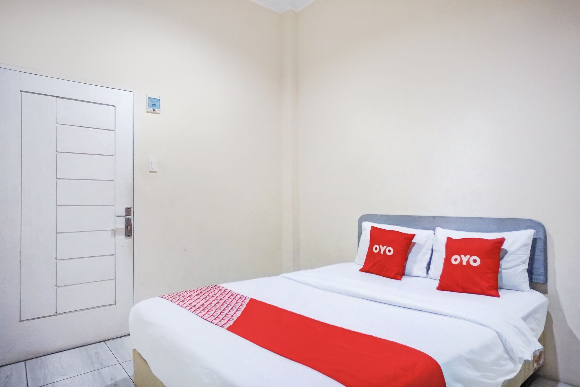 A hotel room with a bed, two pillows with 'OYO' branding, a white door, and a small control panel on the wall.