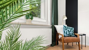 Et værelse med en træstol, et lille bord, en lampe, en potteplante og et vindue med gardiner.