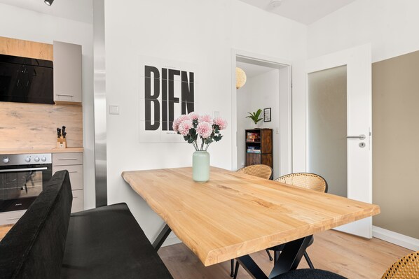 Une cuisine moderne équipée d’un four Juno, un coin repas avec une table et des chaises en bois, et un mur orné d’un panneau indiquant le mot « BIEN ».