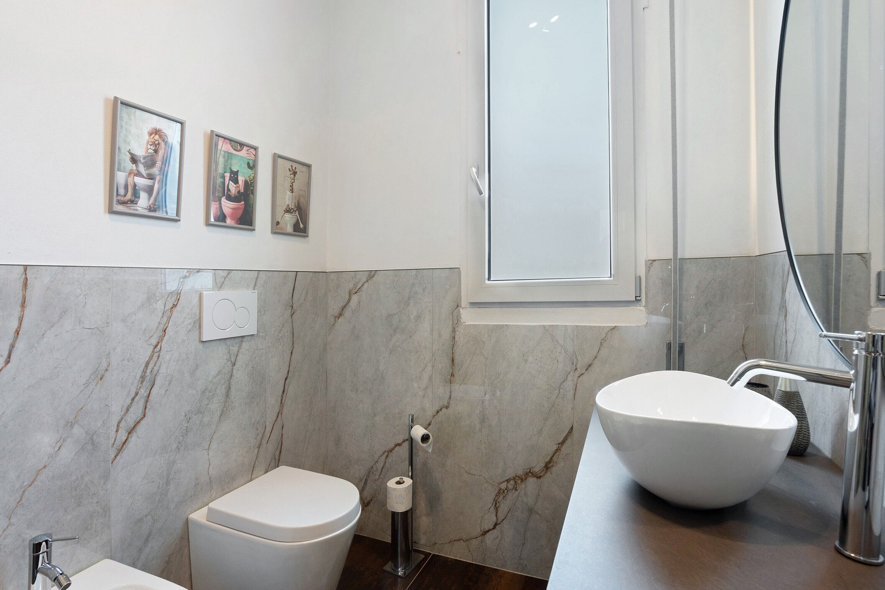 Un baño moderno con inodoro, bidé y lavabo, paredes de mármol y tres obras de arte enmarcadas en la pared.