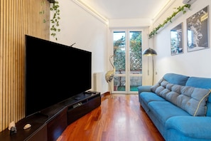 Una sala de estar con televisión de pantalla plana, un sofá azul, piso de madera y vistas a través de un ventanal.