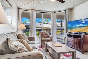 A living room with a sofa, armchair, coffee table, TV, and large windows with a view of palm trees and buildings. - Beautiful 1 BR Oasis Maui Banyan (Kihei)