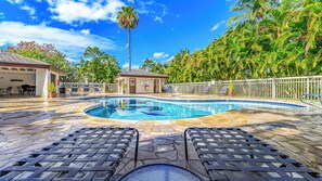 A swimming pool with lounge chairs, a covered seating area, and palm trees in the background.