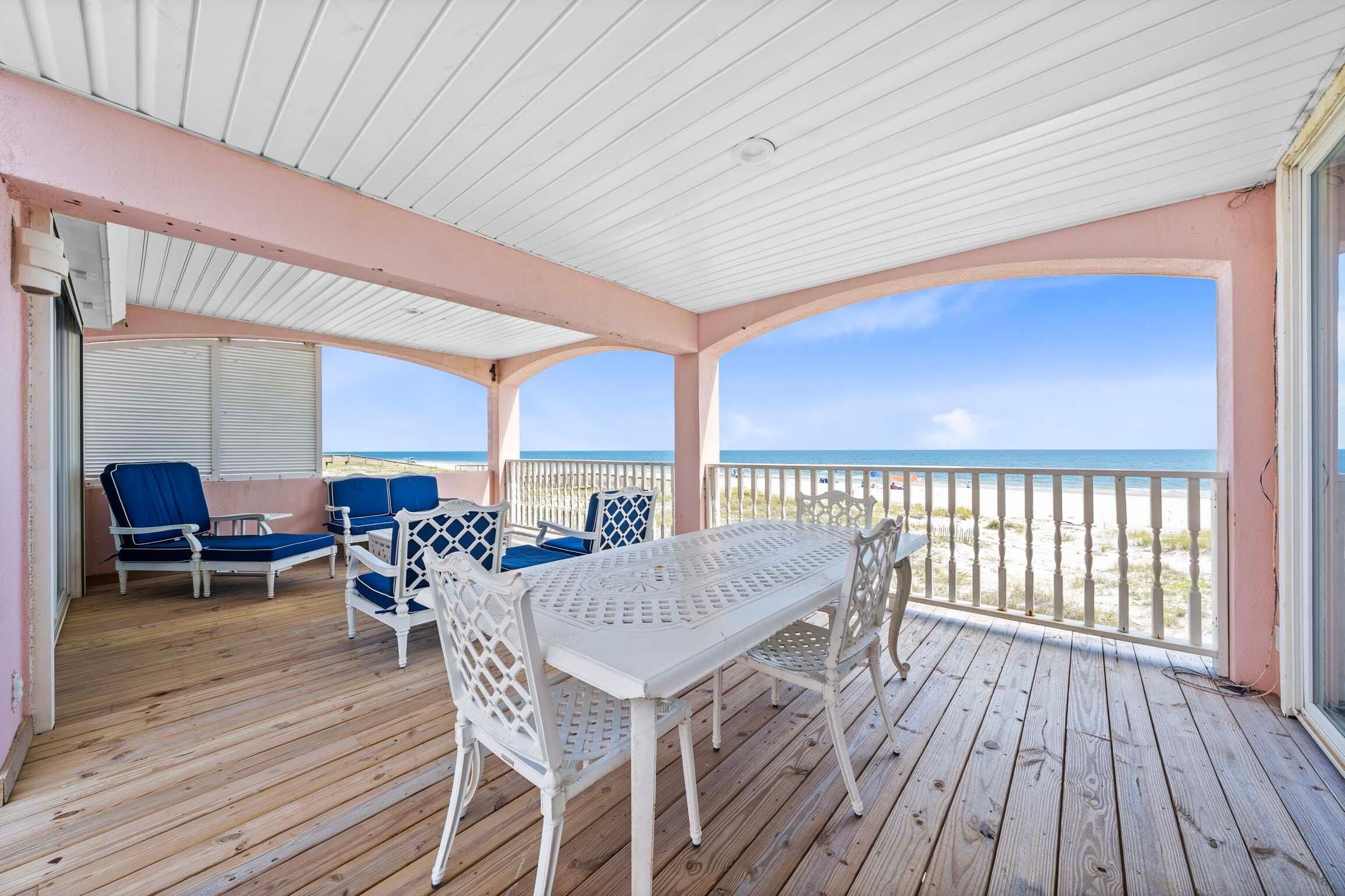 Un espacio al aire libre techado con una mesa y sillas blancas, una silla azul acolchada y vista a la playa.