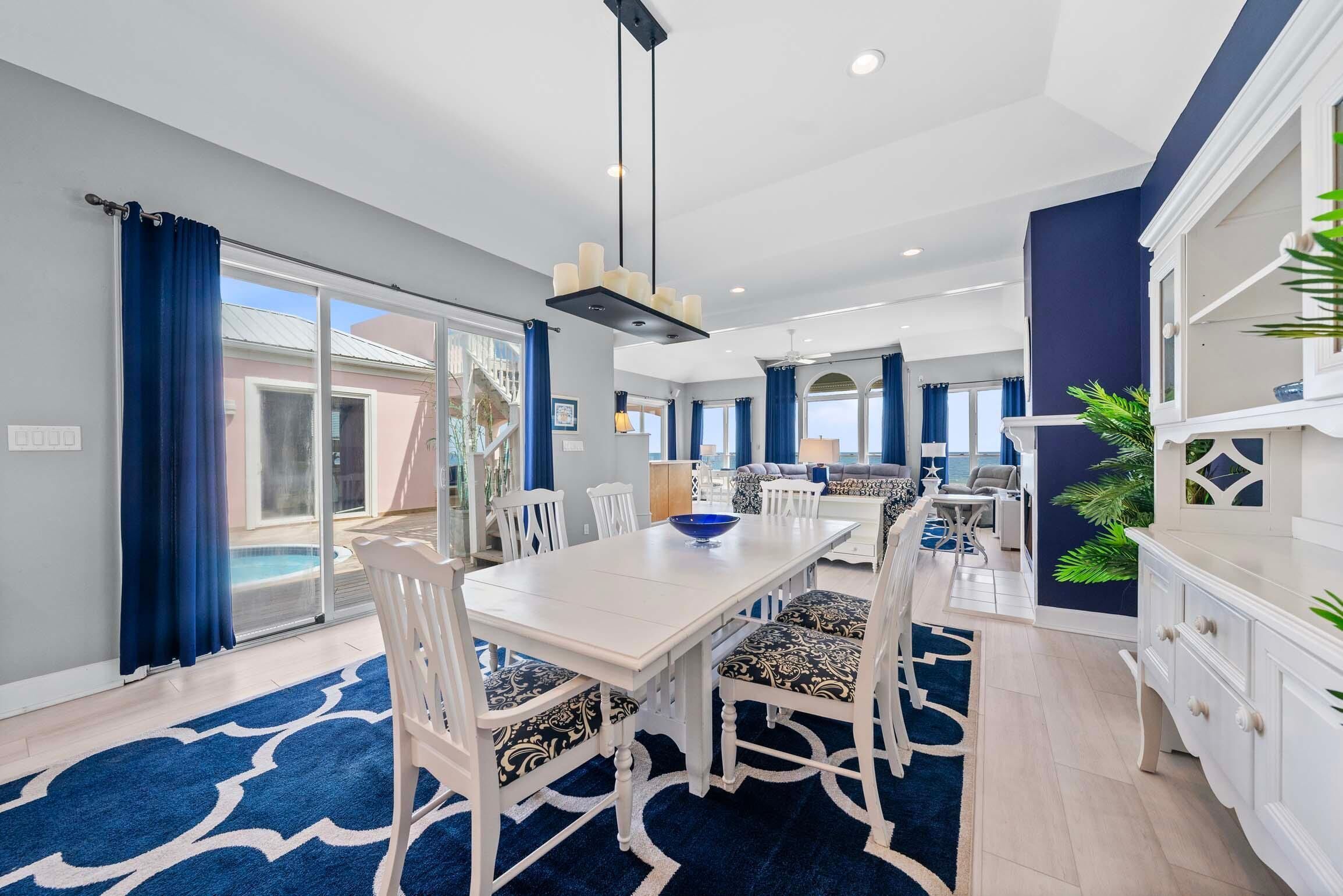 A modern dining area with a white table and chairs, a blue patterned rug, and a large window with blue curtains.
