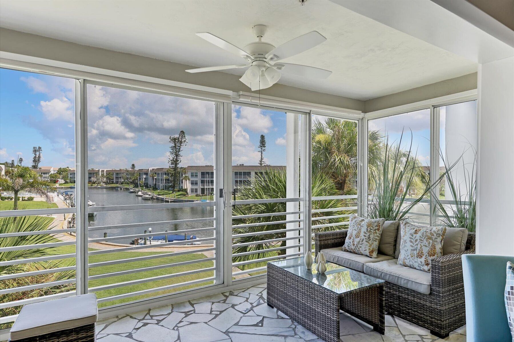 A balcony with wicker furniture, a glass table, and a view of a marina and buildings.