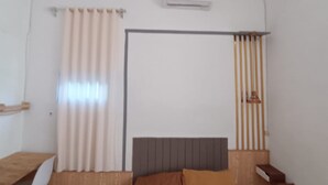 Ein Schlafzimmer mit einem Bett, einem Nachttisch aus Holz, einem kleinen Tisch, einem Fenster mit Vorhängen und einer Wand-Klimaanlage.