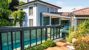 A modern outdoor deck with wooden flooring, two white wireframe tables, and a swimming pool in the background.