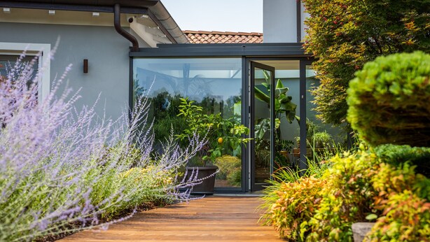 A wooden pathway leading to a modern house with glass doors, surrounded by lush greenery and flowering plants.