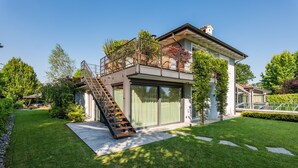 A modern two-story house with a large balcony, a staircase, and a well-maintained lawn.