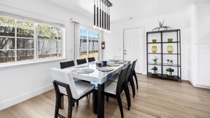A dining area with a white table set for six, black chairs, a modern pendant light, and a plant shelf with decorative items.
