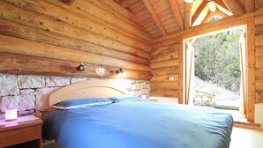 A wooden cabin bedroom with a stone wall, a bed with blue bedding, a bedside table, and a window. - Baita Costa di Gial (Lamon)