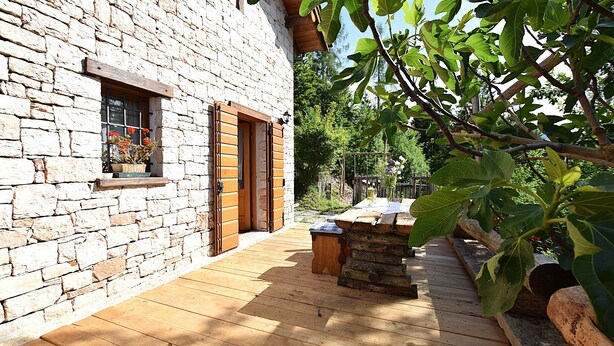 A stone building with a wooden deck, a window with shutters, and a small outdoor seating area. - Baita Costa di Gial (Lamon)