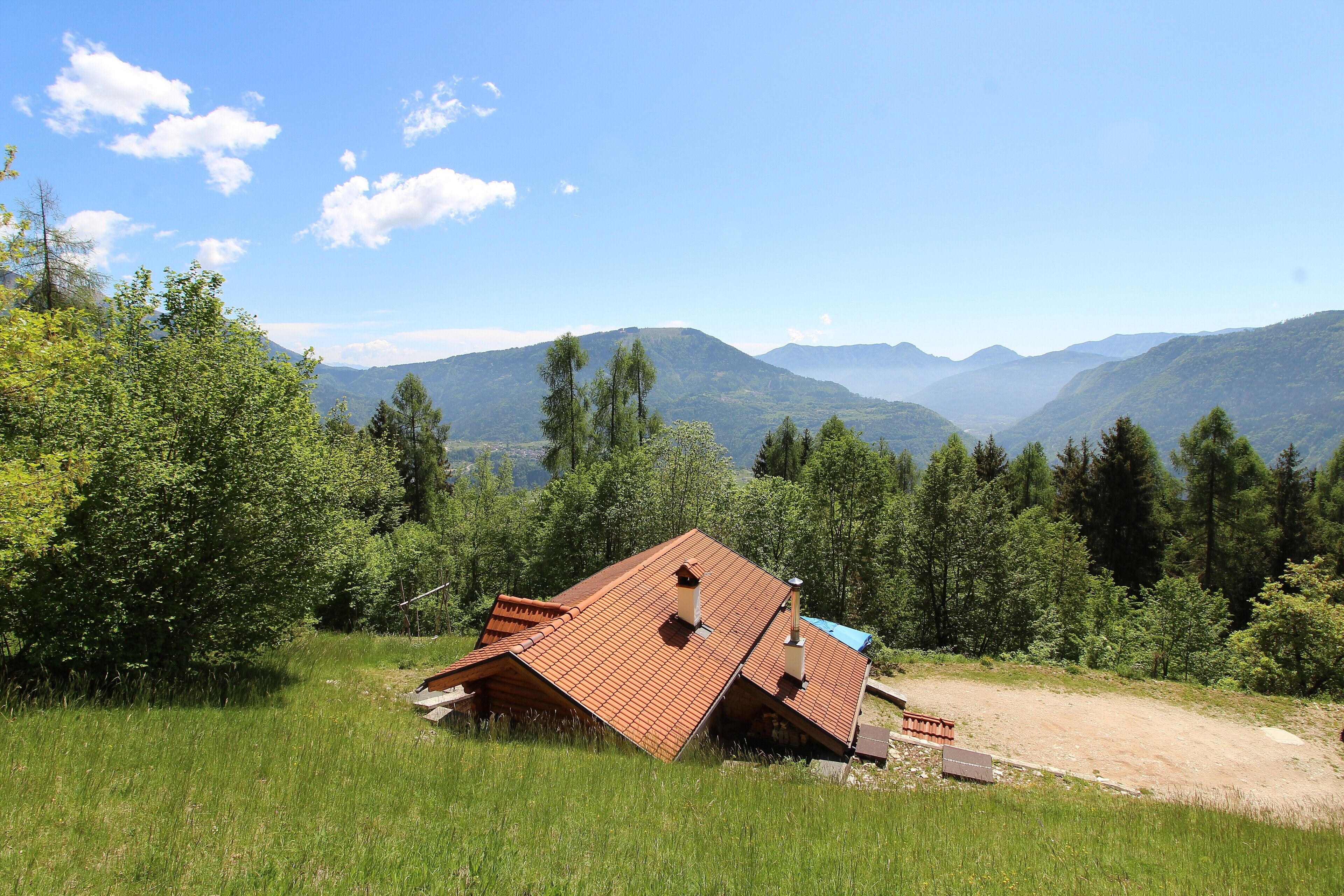 A house with a sloped roof, surrounded by a grassy field and trees, with mountains in the background.