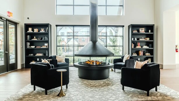 A spacious living room with a central fireplace, black sofas, and a large window offering a view of buildings.