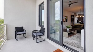 A balcony with two chairs, a glass door leading to an interior space with a ceiling fan, a dining area, and a kitchen. - Modern condo, steps to beach. Gym, generator, gated community. (Nassau)