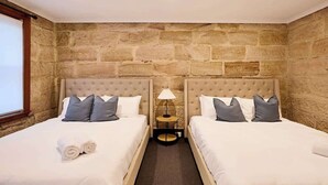 Una habitación con dos camas, paredes de piedra, una mesita con una lámpara y una ventana.