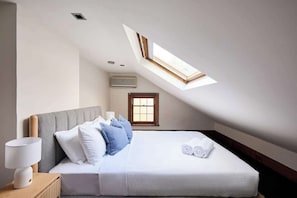 Une chambre avec un puits de lumière, un lit avec une literie blanche, une table de chevet avec une lampe et une fenêtre avec des stores.
