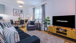 Een moderne woonkamer met een bank, een eethoek met stoelen, een tv op een houten dressoir en een groot raam met gordijnen.