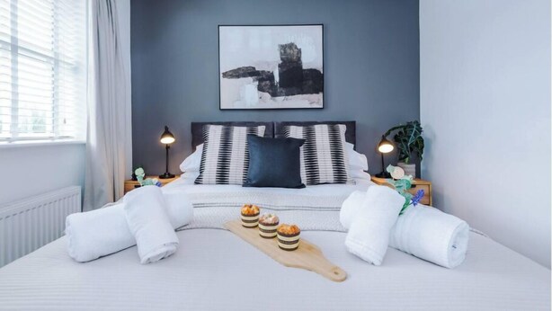 Een net opgemaakt bed met witte lakens, een houten dienblad met gebak en een lijst met een abstract schilderij aan de muur.