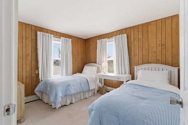 Un dormitorio con dos camas, paredes de paneles de madera, una ventana con cortinas, una mesita de noche con una lámpara y una pequeña mesita de noche con una cesta.