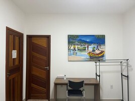 Quarto com porta de madeira, uma escrivaninha com cadeira, pintura de uma praia e um varal de roupas.