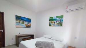 A bedroom with a bed, a small bedside table, a nightstand, two paintings on the walls, and an air conditioning unit. - Suítes Castanheiras  (Paraty)
