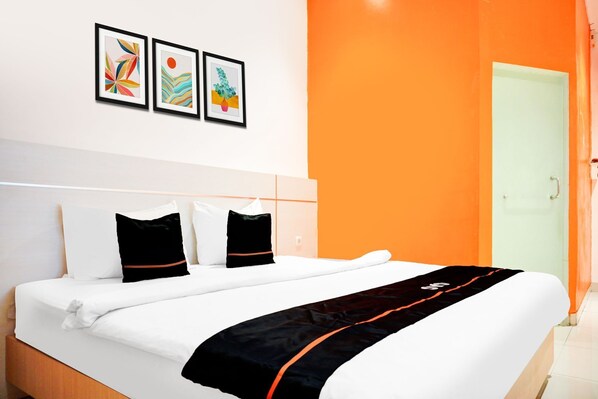 A hotel room with a bed, white bedding, black and orange pillows, a black and orange throw, and three framed artworks on the wall.