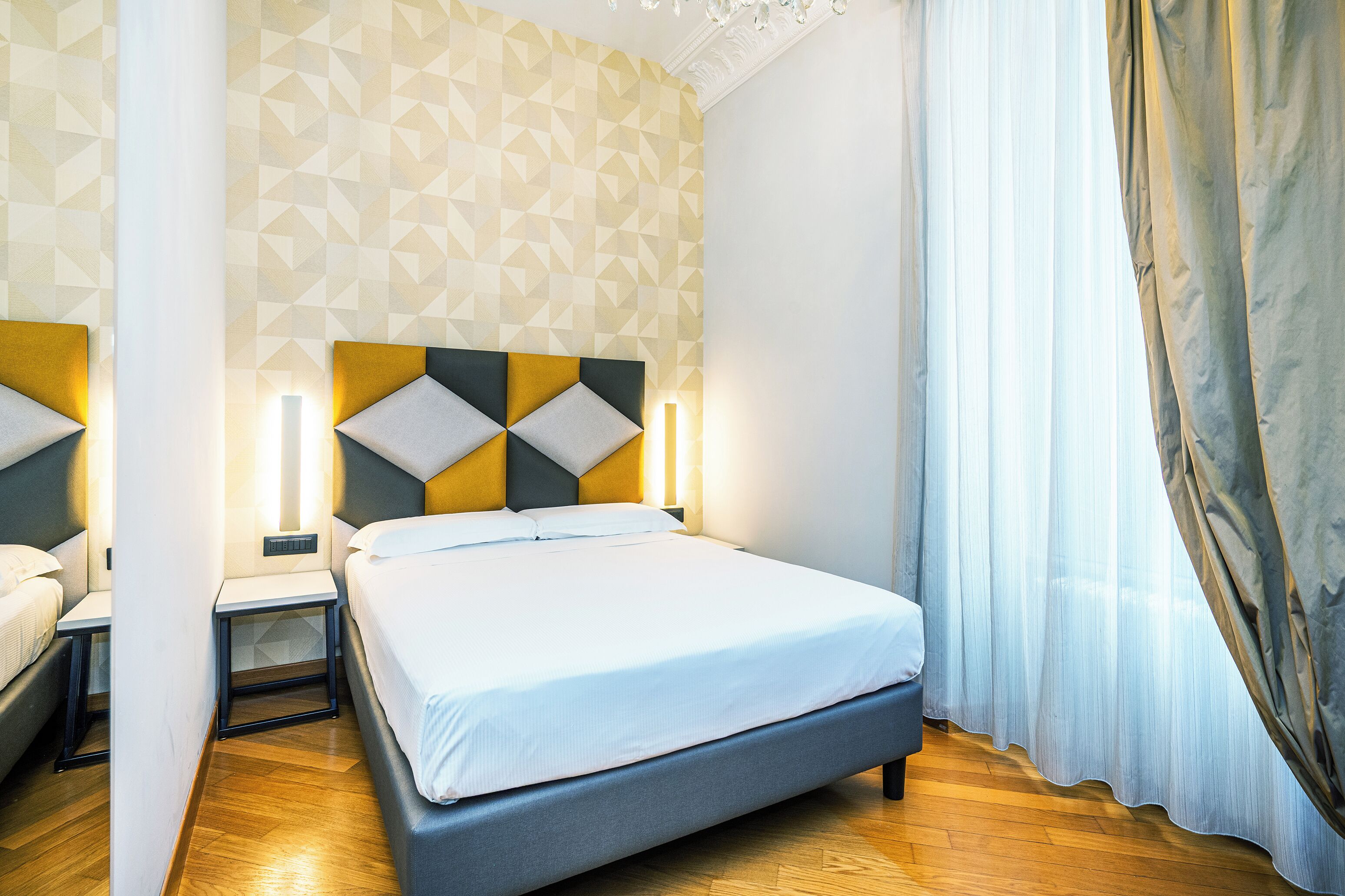 A modern hotel room with a geometric patterned wall, a bed with a grey headboard, bedside tables with lamps, and a wooden floor.