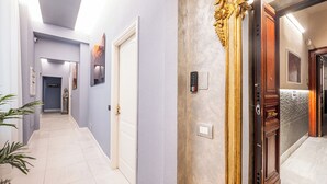 A hallway with a light blue wall, a white door, and a wooden door with a brass handle. There are potted plants on the left side and a light switch on the wall.