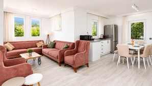 A modern living room with a sofa set, a dining table with chairs, a kitchen area with a refrigerator, and large windows offering a view of greenery. - Daisy Villa - Five Bedroom Villa, Sleeps 10 (Dalyan)