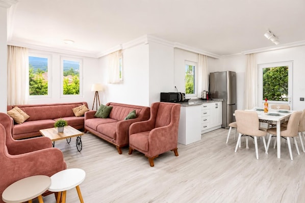 A modern living room with a sofa set, a dining table with chairs, a kitchen area with a refrigerator, and large windows offering a view of greenery.