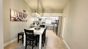 A dining area with a white table and black chairs, a painting on the wall, and a kitchen in the background. - Relaxed Cozy Pool House (Bakersfield)