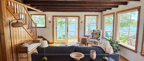 A cozy living room with a dark sofa, wooden floors, a staircase, and large windows offering a view of the outdoors.