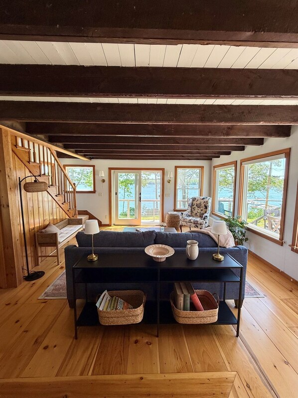 A cozy living room with a dark sofa, wooden floors, a staircase, and large windows offering a view of the outdoors.