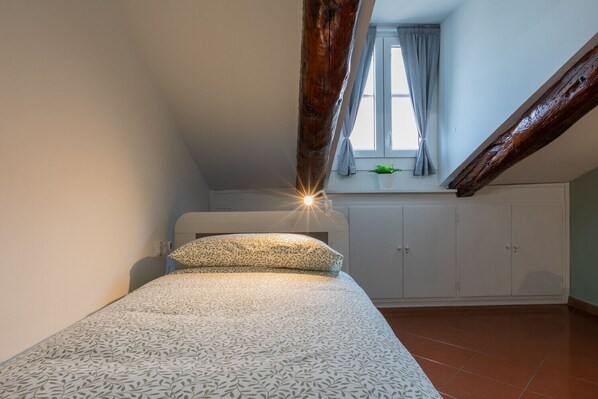 A small, cozy bedroom with a sloped ceiling, a bed with patterned bedding, a window with curtains, and a wooden beam.