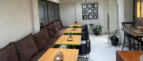 Un espace repas moderne avec de longues tables en bois et des chaises noires, un canapé marron et des plantes en pot.