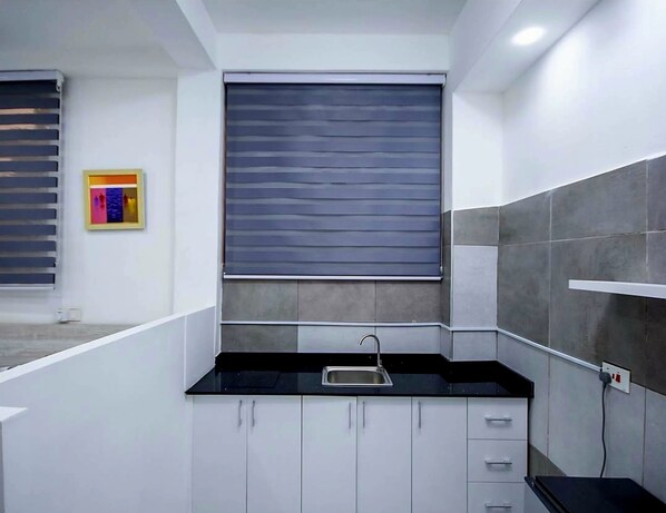 A modern kitchen with white cabinets, a black countertop, and a window with horizontal blinds. - Milton Avenue Suites (Accra)