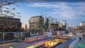 A rooftop lounge with seating areas and a fire feature, surrounded by city buildings.