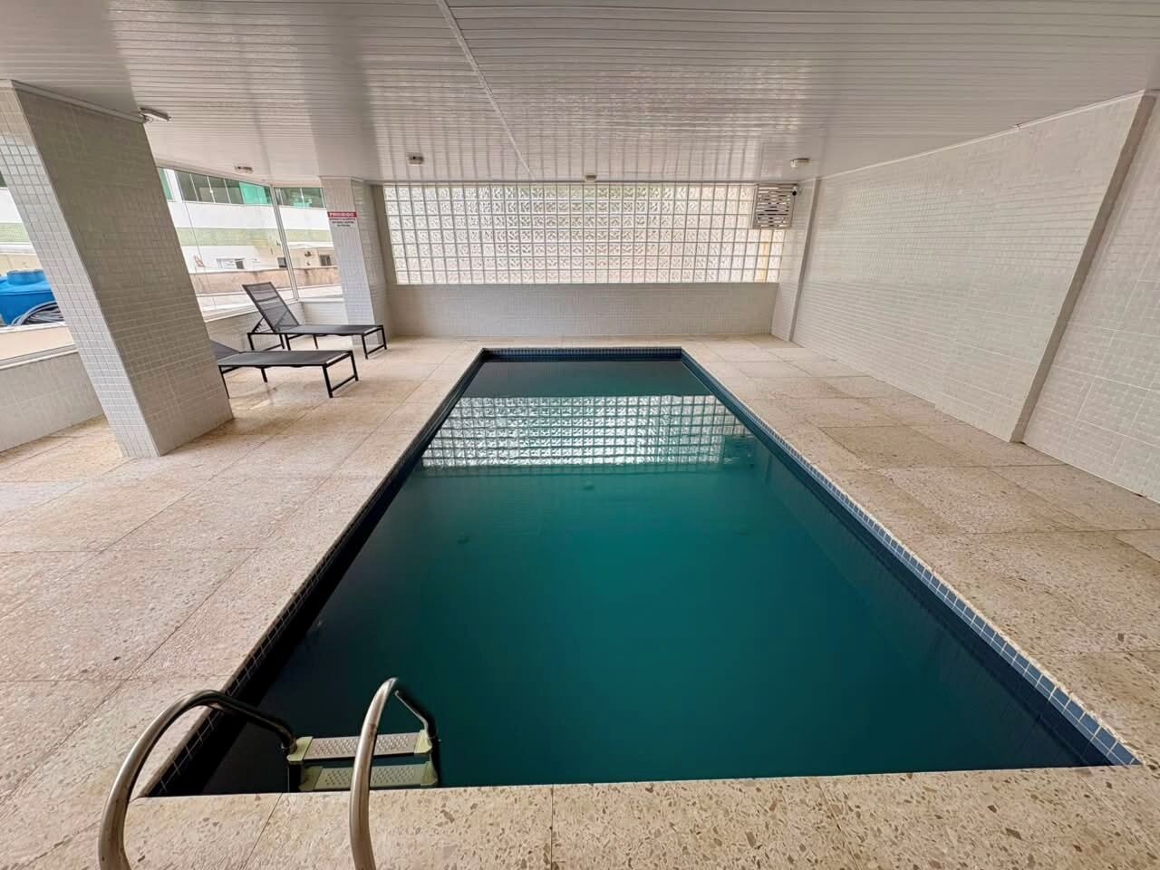 Ein Schwimmbecken mit gefliestem Sonnendeck, einer Leiter und einem überdachten Bereich mit Sitzgelegenheiten.